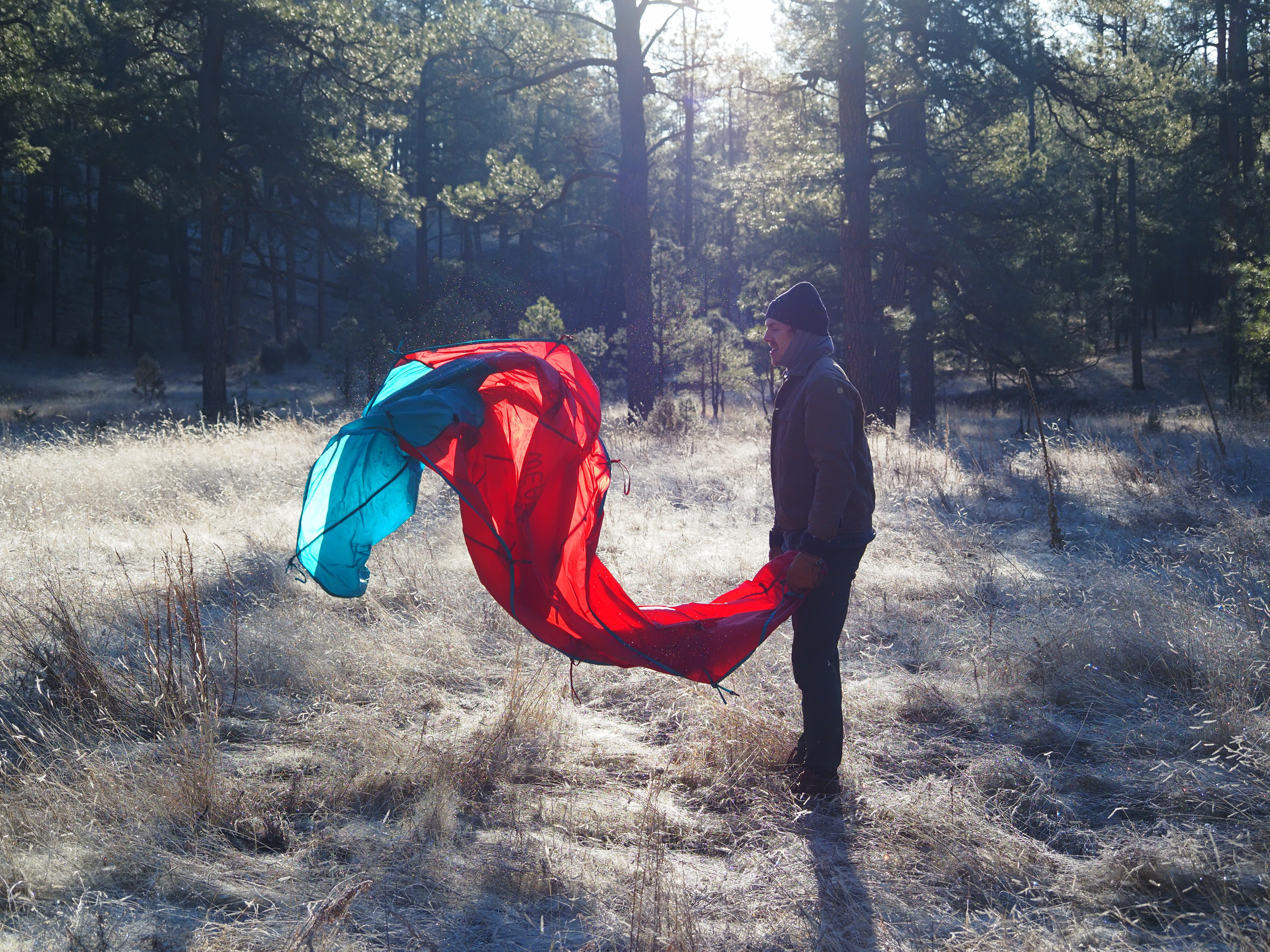 A person shaking out a tent in a frosty forest lit by morning sun