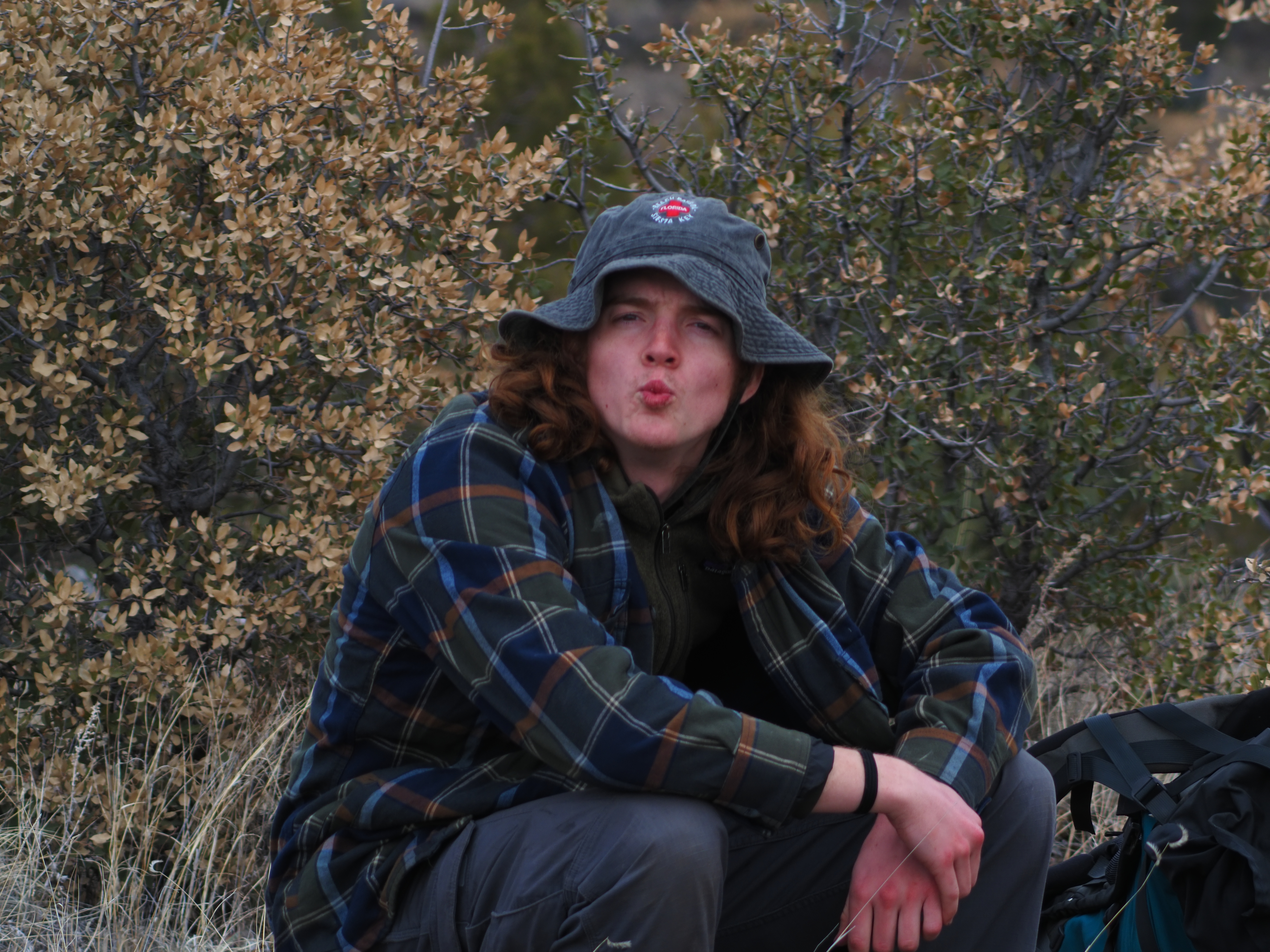 A hiker in a bucket hat and plaid flannel sitting next to a backpack on the trail