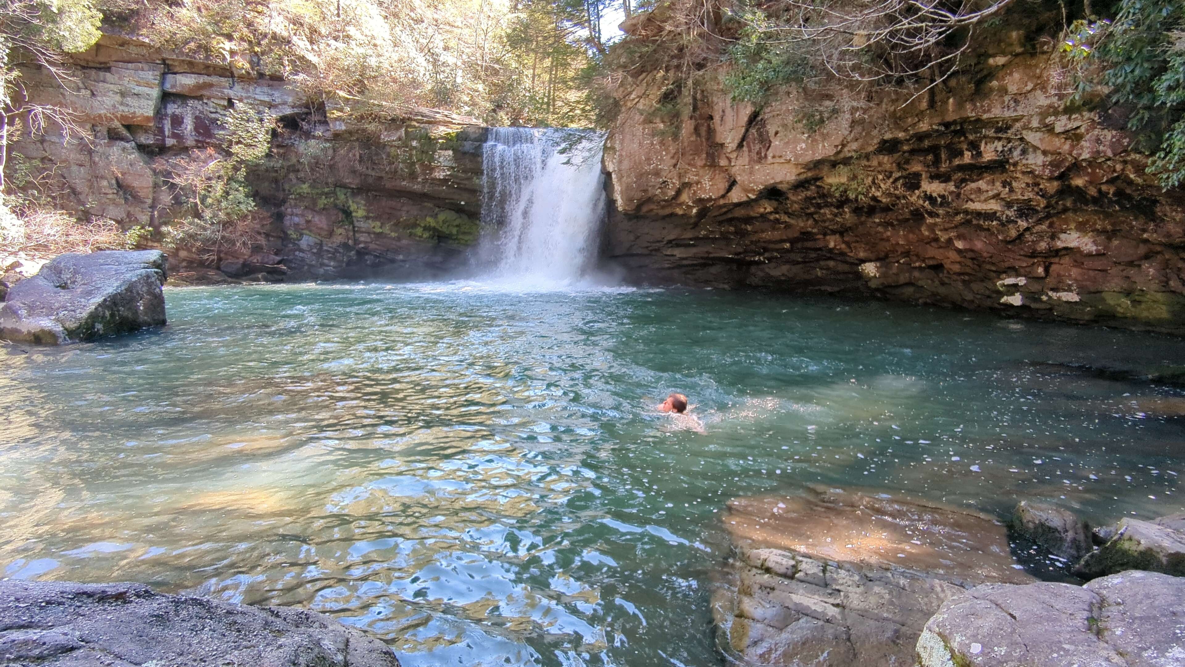 A guy swimming in a pool of water at the bottom of a waterfall